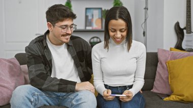 Evlerindeki koltukta oturan gülümseyen bir çift hamilelik testi sonucuna mutlu bir şekilde bakıyor..