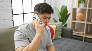 Genç Asyalı bir adam modern bir oturma odasında akıllı bir telefonla konuşuyor, gelişigüzel, bağlı bir yaşam tarzını yansıtıyor..