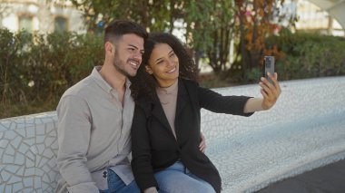 Gülümseyen bir çift parktaki bankta selfie çeker ve şehir ortamında sevgilerini ve mutluluklarını sergilerler..