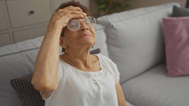 Kısa saçlı ve gözlüklü yaşlı İspanyol kadın evinin oturma odasında otururken alnına dokunuyor..