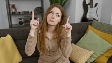 Düşünceli genç sarışın bir kadın, rahat bir oturma odasında gri bir kanepede otururken yukarı doğru hareket eder..