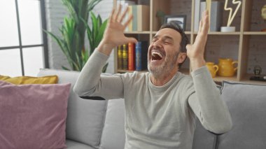 Orta yaşlı bir adam, rahat bir oturma odasında kitap ve dekor dolu bir koltukta dinleniyor..