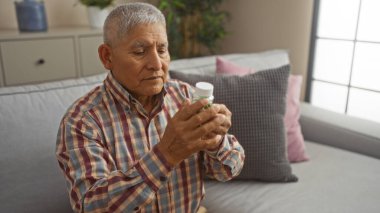 Gri saçlı İspanyol bir adam, gri ve pembe yastıklı, odaklanmış bir ifadeyle modern bir oturma odasında otururken ilaç şişesini inceliyor..