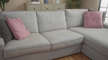 Rahat ve modern oturma odasının içinde pembe yastıklı gri bir kanepe, ahşap döşeme, kapalı salonda bitkiler ve dekoratif eşyalarla dolu bir dolap var..