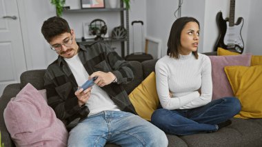 Bir erkek ve bir kadın koltuğa oturur, erkek akıllı telefonuna odaklanır, kadın ise sinirli görünür, modern bir oturma odasında..