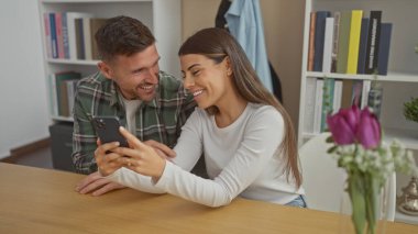 Sırıtkan bir çift ahşap bir masada kitaplığı olan rahat bir oturma odasında akıllı telefon kullanıyor..