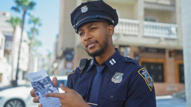 Afrika kökenli Amerikalı bir polis memuru güneşli bir caddede kayıp ilanı tutuyor.