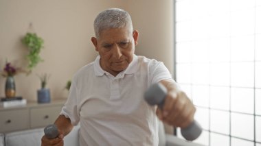 Beyaz saçlı İspanyol bir adam modern bir oturma odasında dambıllarla egzersiz yapıyor, rahat bir ev ortamında sağlık ve sağlığın somutlaştığı bir yer..
