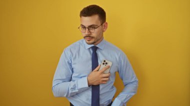 Sakallı yakışıklı, genç, İspanyol bir adam sarı arka planda akıllı bir telefon tutuyor, mavi gömlek ve kravat takmış..