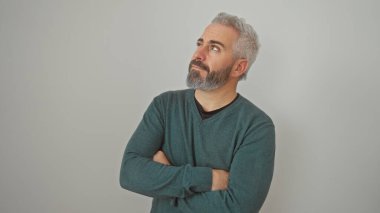 Düşünceli, sakallı ve gri saçlı bir adam beyaz bir arka plan karşısında duruyor, kollarını kavuşturmuş, başka tarafa bakıyor..