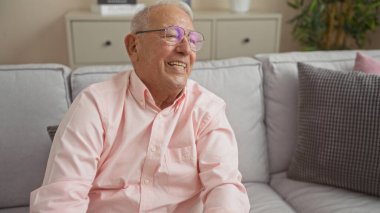Beyaz saçlı, gözlüklü, rahat bir oturma odasında oturmuş gülümseyen yaşlı, beyaz bir adam..