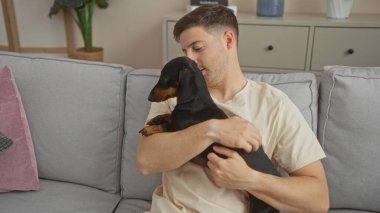 Sıcak bir oturma odasında kanepede otururken elinde daksund köpeği tutan genç bir İspanyol..