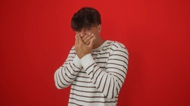 Genç, yakışıklı İspanyol bir adamın portresi çizgili süveteri ile izole edilmiş kırmızı arka plan duvarının üzerinde poz veriyor, elleriyle ağzını kapatıyor..