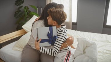 Bir kadın ve oğlu bir yatakta sevgi dolu bir kucaklaşmayı paylaşıyor, bir hediye tutan çocukla, evde şefkatli bir aile anını tasvir ediyor..