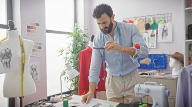 Odaklanmış orta yaşlı sakallı bir adam renkli bir terzi dükkanında tasarım çizimleri ve bir mankenle fotoğraf çekiyor..