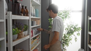 Ev dekorasyonu dükkanında sakallı genç bir adam. Raflardaki çeşitli dekoratif ürünlerle dolu şeyleri inceliyor. Yumuşak doğal ışık altında büyük bir ev bitkisinin yanında..