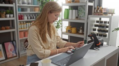 Ev dekorasyonu mağazasında çalışan genç bir kadın. Sarı saçlı, dizüstü bilgisayar ve hesap makinesi kullanan, çeşitli dekoratif malzemelerle çevrili.