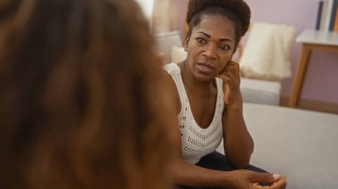 Kadın, sıcak bir evin oturma odasında arkadaşlarıyla konuşuyor. Oturmuş ve derin bir sohbete dalmış. Kaygı ve empati ifadeleri sergiliyor..