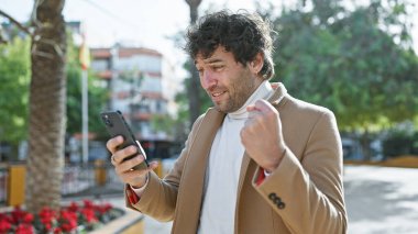 Güneşli bir parkta, yeşil ve çiçeklerle çevrili telefonuna gülümseyen sakallı yakışıklı bir İspanyol..