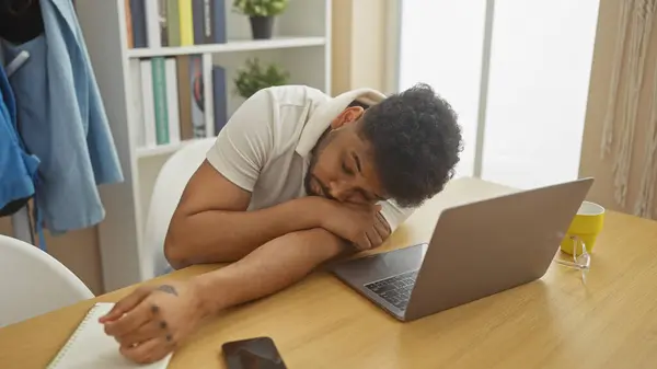 stock image Tired african american man napping at his home desk with a laptop, phone, and glasses nearby.