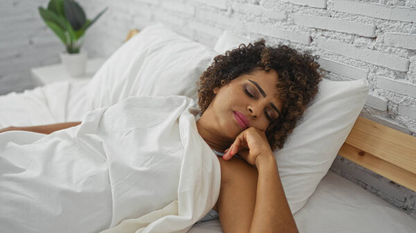 Красивая женщина с вьющимися волосами мирно спит на кровати в уютной спальне с белыми кирпичными стенами и внутренним декором растений, предполагая спокойную домашнюю обстановку.