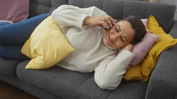 stock image A middle-aged hispanic woman relaxes on a couch at home, talking on the phone with a calm expression.