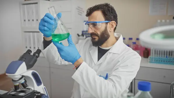 stock image A bearded scientist man examines green liquid in flask at a laboratory, surrounded by scientific equipment and wearing protective glasses and gloves.