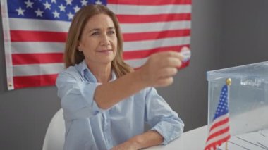 Orta yaşlı bir kadın arka planda Amerikan bayrağıyla kapalı alanda oy veriyor. Demokrasi ve vatandaşlık taşıyor..