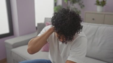 Genç İspanyol bir adam kanepede otururken boyun ağrısı çekiyor. Rahatsızlık ve stres gösteriyor..