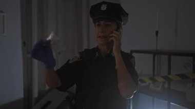 Orta yaşlı bir kadın polis telefonda konuşurken ve elinde kanıt tutarken kapalı bir suç mahallini araştırıyor..