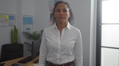 Ciddi orta yaşlı bir kadın işyerinde odaklanmış ifadeyle kollarını kavuşturuyor..