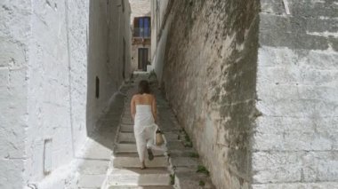 Beyaz elbiseli genç bir İspanyol kadın, antik taş duvarlar ve dar geçitlerle çevrili eski bir kasaba olan Ostuni, Puglia, İtalya, Avrupa 'da taş basamaklardan yukarı yürüyor..