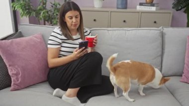 Genç bir kadın oturma odasında rahat bir şekilde oturuyor. Kanepede Chihuahua köpeği eşliğinde telefonunu kullanırken kırmızı bardaktan yudumluyor..