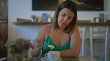 Genç bir kadın kahvesini karıştırıyor, bir İtalyan kafeteryasında oturuyor, sakin bir ortam ve modern bir dekorla..