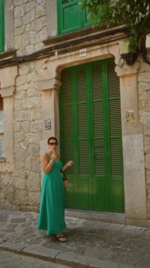 Yeşil elbiseli genç bir kadın Valldemossa, Mallorca 'da dondurma yiyor. Taş bir binanın önünde yeşil kepenkleri olan bir kadın.