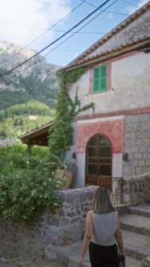 Mallorca, İspanya 'nın pitoresk taş caddelerini keşfeden bir kadın. Arka planda, yemyeşil dağlar ve açık gökyüzüyle çevrili geleneksel bir kasaba evi var.