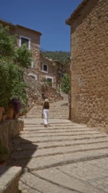 Güneşli, pitoresk bir köy olan Mallorca, İspanya 'da gezen, şık bir yaz elbisesi ve güneş gözlüğü takan kırsal taştan binalar ve yemyeşil yeşilliklerle çevrili bir kadın..
