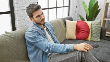 Modern bir oturma odasının kumandalı kanepesinde uzanan sıradan bir kot ceketli yakışıklı bir İspanyol..