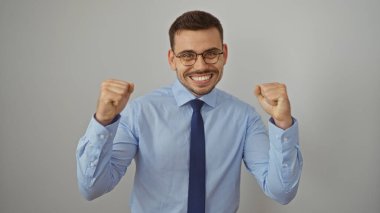 Gözlüklü ve mavi gömlekli yakışıklı genç adam, izole edilmiş beyaz bir arka plana yumruk atarak kutlama yapıyor..