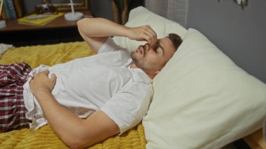 Sakallı, beyaz tişört ve ekose pijama pantolon giymiş, yatak odasında rahat değilmiş gibi görünen, yatak odasındaki bir yatağa uzanmış genç bir İspanyol..