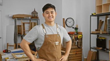 Kendine güvenen genç bir Asyalı iyi organize edilmiş bir marangozluk atölyesinde gururla duruyor, profesyonelliği canlandırıyor..
