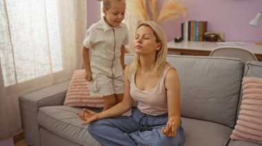 Anne ve kızı sıcak bir oturma odasında birlikte vakit geçiriyorlar, çocuk yanında dururken, kadın kanepede meditasyon yapıyor, evde huzurlu bir anın tadını çıkarıyor.