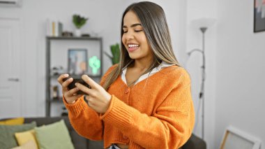 Modern bir oturma odasında akıllı telefon kullanarak şık turuncu kazaklı, gülümseyen İspanyol kadın..