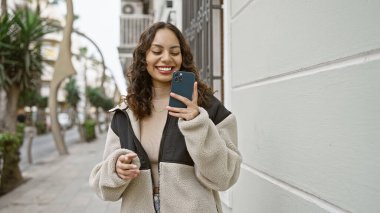 Bir şehir caddesinde yürürken akıllı telefon kullanan gülümseyen genç bir kadın, kentsel bağlantı ve günlük yaşam tarzını aktarıyor..