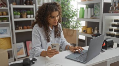 Dizüstü bilgisayarda çalışan ve arka planda dekoratif eşya rafları olan bir mağazada akıllı telefon kullanan bir kadın.