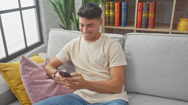 Kanepede akıllı telefon kullanırken gülümseyen yakışıklı İspanyol adam.