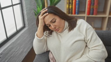 Orta yaşlı bir kadın rahat bir oturma odasında stresli hissediyor. Endişe ya da baş ağrısı göstergesi olarak alnına dokunuyor..