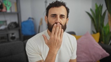 Ağzını kapatan sakallı orta yaşlı bir adam modern bir oturma odasında sürpriz ya da sessizlik jesti sergiliyor..