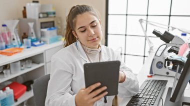 Genç bir İspanyol kadın bilim adamı, teknoloji ve sağlık hizmetlerini temsil eden bir laboratuvar ortamında bir tablet üzerindeki verileri analiz ediyor..