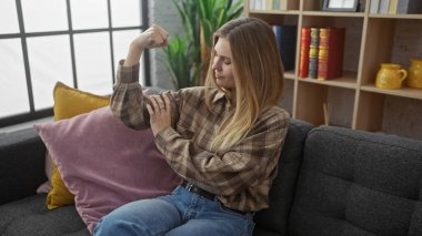 Blonde woman flexing arm muscles indoors, portraying strength on a cozy living room couch setting. clipart